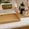 A person holding a natural rattan and bamboo serving tray set with coffee mugs and a French press
