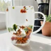 A double-walled glass mug with real preserved dried flowers inside the walls, displayed on a table with a plant and coffee machine in the background