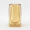 A Double Glass Mate Gourd – 100ml Insulated Yerba Cup, featuring a sleek, double-walled glass design. The cup has a square-shaped clear outer wall and a rounded, organic inner bulb. The entire glass structure is tinted with a warm, transparent amber honey color, resting against a clean, minimal white background.